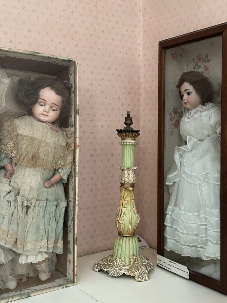 Zwei Puppen: rechts eine in einer Glasvitrine, links eine in einem blumengemusterten Karton, der an eine Kinderlade oder einen Sarg erinnert. Zwischen ihnen steht eine grün-goldene Tischlampe im floralen Stil. Aufgenommen in einem inszenierten viktorianischen Kinderzimmer – ein Bild wie aus einem eingefrorenen Traum.