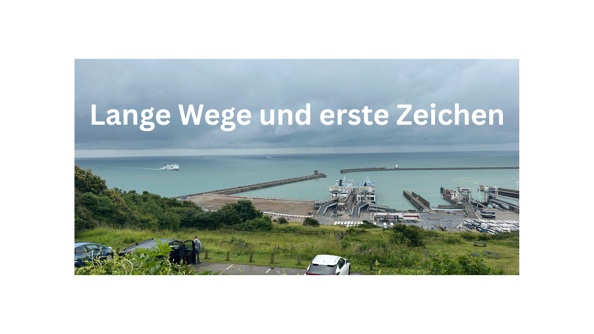 Blick auf den Hafen von Dover. Im Vordergrund ein Parkplatz, im Hintergrund das Meer, die Fähren und das Tor nach England. Das Bild trägt den Titel: "Lange Wege und erste Zeichen".