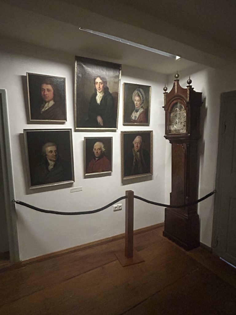 Wand im Gleimhaus Halberstadt mit historischen Portraitgemälden aus dem 18. Jahrhundert. Zu sehen sind sechs gerahmte Bilder von Gleims Zeitgenossen sowie eine antike Standuhr. Ein Absperrseil markiert den Abstand zum Ausstellungsbereich.