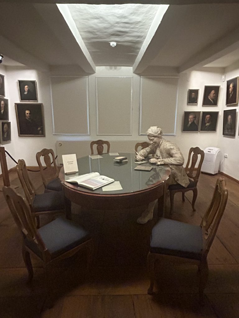 Innenansicht des „Tempels der Freundschaft“ im Gleimhaus Halberstadt: Ein ovaler Tisch umgeben von leeren Stühlen, darauf Bücher und Dokumente. Am Tisch sitzt eine lebensgroße Figur Johann Wilhelm Ludwig Gleims, als stiller Gastgeber unter Portraits seiner Weggefährten.
